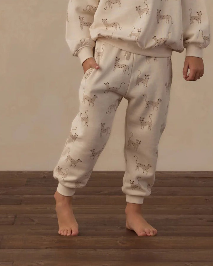 A toddler stands barefoot on a wooden floor wearing a cream-colored two-piece pajama set with a light brown deer print, paired with a rust-red knit beanie. The background features a textured beige wall with decorative leaf-like cutouts.
